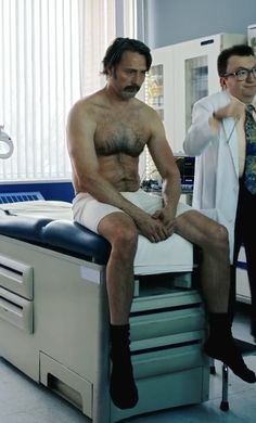 a shirtless man sitting on top of a hospital bed next to another man in a white coat