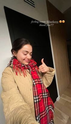 a woman in a tan jacket and red scarf giving the thumbs up sign with her hand