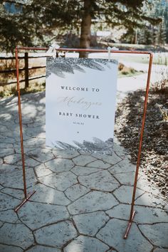 a welcome sign for a baby shower is displayed on a metal stand in front of a tree