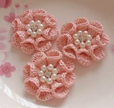 three pink crocheted flowers with pearls on them sitting on a plate next to some flowers