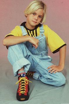 a young boy sitting on the ground wearing overalls and yellow t - shirt with red shoes