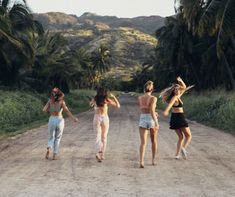 four girls walking down a dirt road in front of palm trees and mountains, with one girl holding her hair back