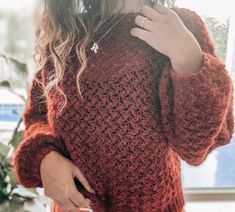 a woman is wearing a red sweater and holding her hand on her shoulder as she stands in front of a window