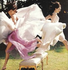 two women in white dresses are jumping over a bench on the grass with their legs spread out