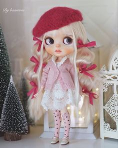 a doll with long blonde hair wearing a red hat and white dress standing in front of a christmas tree