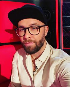 a man wearing glasses and a hat sitting in front of a red wall with speakers