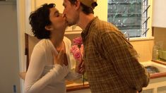 a man and woman kissing each other in front of a kitchen sink with flowers on the counter