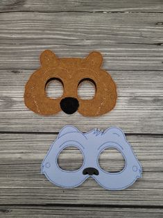 two masks are sitting next to each other on a wooden surface, one has a bear face and the other has a blue mask