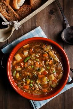 a bowl of vegetable soup next to some bread and spoons on a wooden table