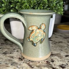 a green ceramic mug with an image of a turtle on the side sitting on a counter next to a potted plant