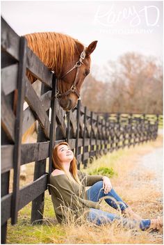 a woman sitting on the ground next to a fence with a horse sticking its head over it