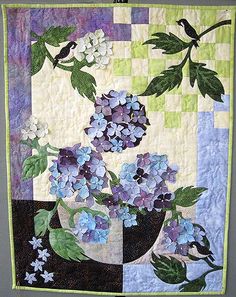 a quilted wall hanging with blue and purple flowers in a vase on a table