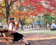 a woman sitting on a wooden bench drinking from a cup in the park with fall leaves around her