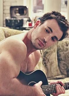 a shirtless man sitting on a couch holding a guitar and looking at the camera