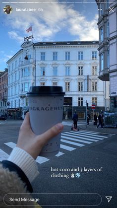 a person holding up a cup of coffee in front of a building with people walking on the street