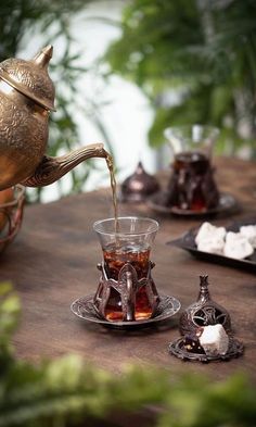 tea being poured into a glass cup on top of a wooden table surrounded by plants