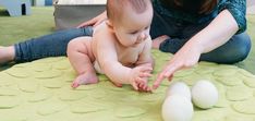 a woman kneeling down next to a baby on top of a green mat with two eggs