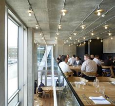 people sitting at tables in a restaurant with large windows and lights on the ceiling above them