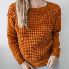 a woman with blonde hair wearing an orange knitted sweater and jeans, standing in front of a white wall