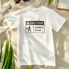 a white t - shirt that says caution student driver on it next to sunglasses and palm leaves