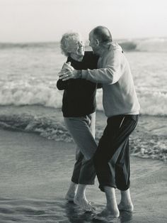 an older couple dancing on the beach in front of the ocean with their arms around each other