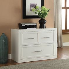 a white dresser with two drawers in front of a window and a painting on the wall