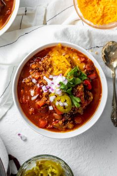 two bowls of chili and cheese soup on a white table with spoons next to it