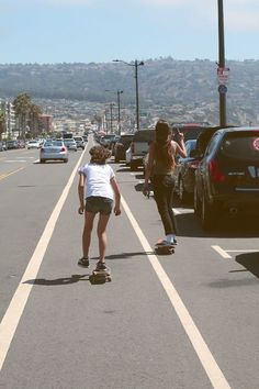 two people riding skateboards down the middle of a street with cars parked on both sides