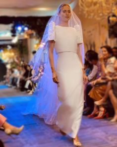 a model walks down the runway in a white dress and veil, with people sitting on the floor watching