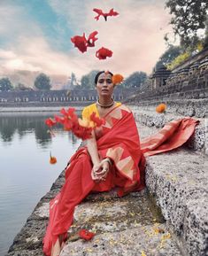 a woman sitting on the edge of a body of water with red flowers in her hair