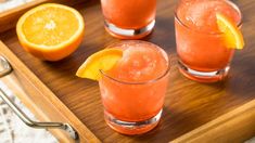 three glasses filled with orange juice and garnished with an orange slice on a wooden tray