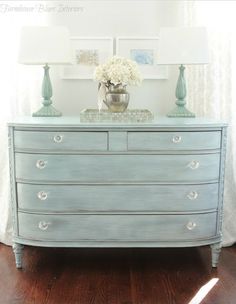 a blue dresser with white flowers on top and two lamps on the side, in front of a window