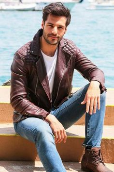 a man sitting on steps next to water wearing a brown leather jacket and blue jeans