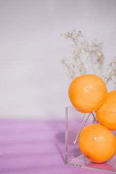 three oranges sitting in a clear vase on a purple tablecloth with white baby's breath flowers