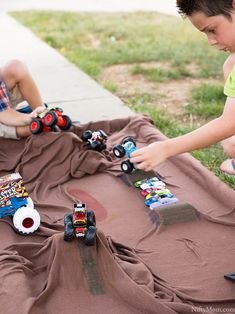 two young boys playing with toy trucks on a blanket in front of a sidewalk and grass