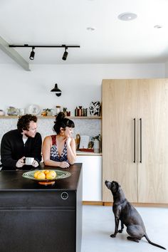 a man and woman sitting at a kitchen counter with a dog standing next to them