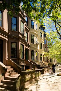 people are walking down the sidewalk in front of some buildings on a sunny day with green trees