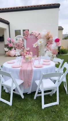 a table set up with white chairs and pink flowers on the top, in front of a house