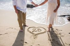 a man and woman holding hands while standing in the sand with a heart drawn on it