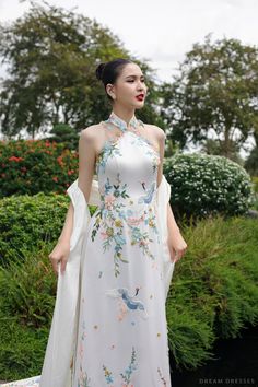 a woman wearing a white dress with flowers on it, standing in front of bushes