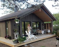 a woman standing on the back porch of a small wooden cabin with patio furniture and potted plants