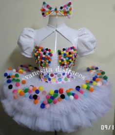 a white dress with multicolored beads and bows on the top is hanging in front of a wall