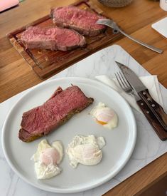 a white plate topped with meat and eggs on top of a wooden table next to utensils