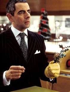 a man wearing a suit and tie holding a flower in his right hand while standing next to a christmas tree