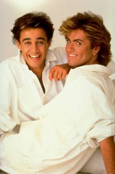 two young men sitting next to each other on top of a white sheet covered bed