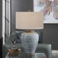 a table lamp sitting on top of a glass table in front of a couch and painting