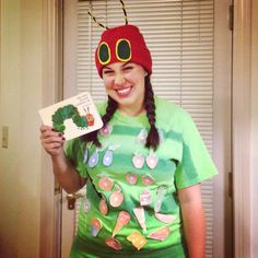 a woman wearing a green shirt and red hat holding up a book with the very hungry caterpillar on it