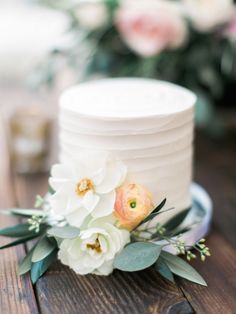 a white wedding cake with flowers on the top is sitting on a wooden table next to some greenery