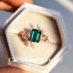 someone is holding an emerald and diamond ring in a white velvet box with its lid open