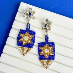 Add a touch of festive charm to your ensemble with these classic Dreidel Beaded Dangle Earrings. Intricately designed with shimmering blue and gold beads, they capture the season's spirit while adding a glamorous sparkle. These earrings are perfect for making a stylish statement at any holiday gathering. Measures 2.5" in length Beaded Star of David Dreidel motif Traditional Diwali Earrings With Dangling Beads, Festive Blue Earrings With Dangling Beads, Blue Beaded Brass Dangle Earrings, Blue Spiritual Dangle Earrings, Blue Hand-strung Dangle Beaded Earrings, Gay Pride Gifts, Shabbat Candles, Bat Mitzvah Gifts, Jewish Jewelry